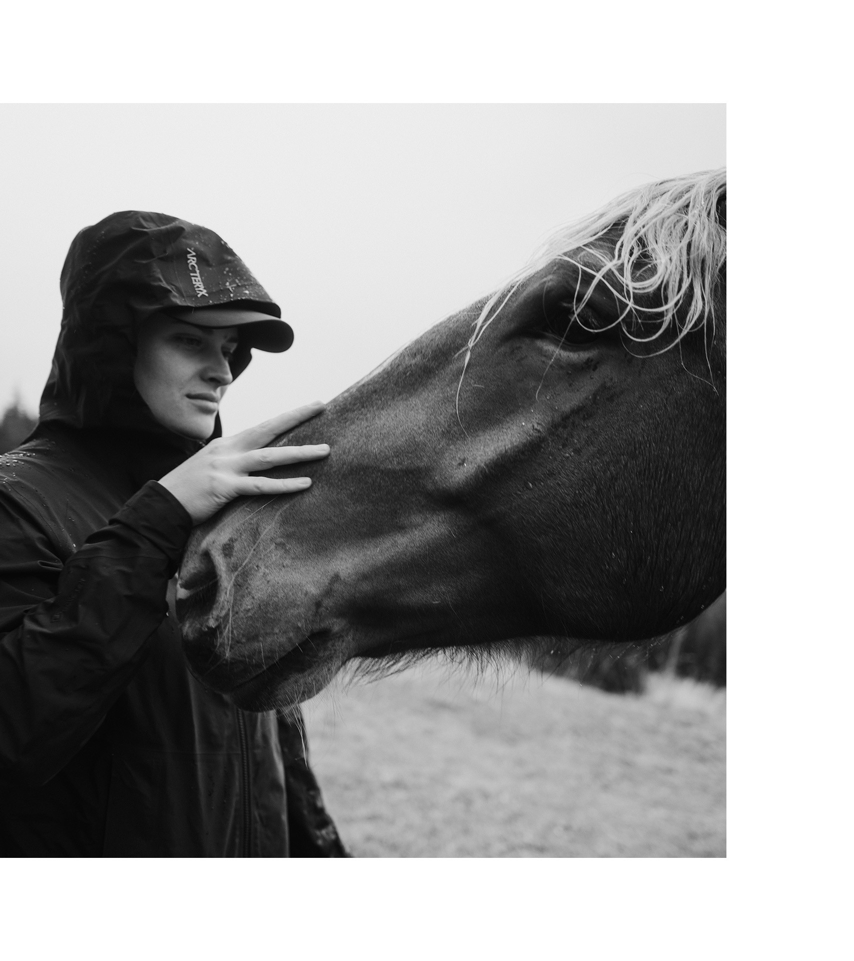 Black and white photograph of Vali Höll stroking a horse while wearing a hooded raincoat.